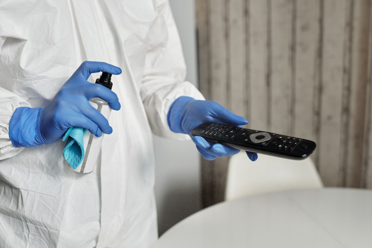 heros-img Hands in gloves disinfecting a remote control indoors. Pandemic hygiene concept.